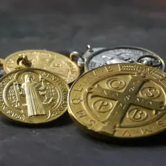 Kilka złotych i srebrnych medalików św. Benedykta leży na ciemnym tle.