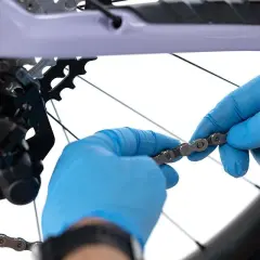 Ręce w niebieskich rękawiczkach manipulują łańcuchem rowerowym, przygotowując go do skrócenia.