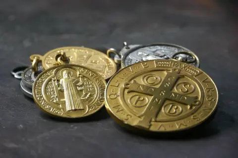 Kilka złotych i srebrnych medalików św. Benedykta leży na ciemnym tle.