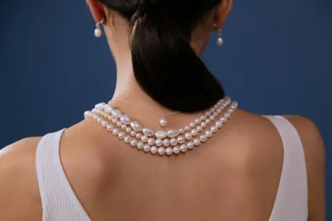 Kobieta z tyłu w białej bluzce, ozdobiona kilkoma naszyjnikami z pereł i kolczykami. Miara pereł podkreśla jej elegancję.