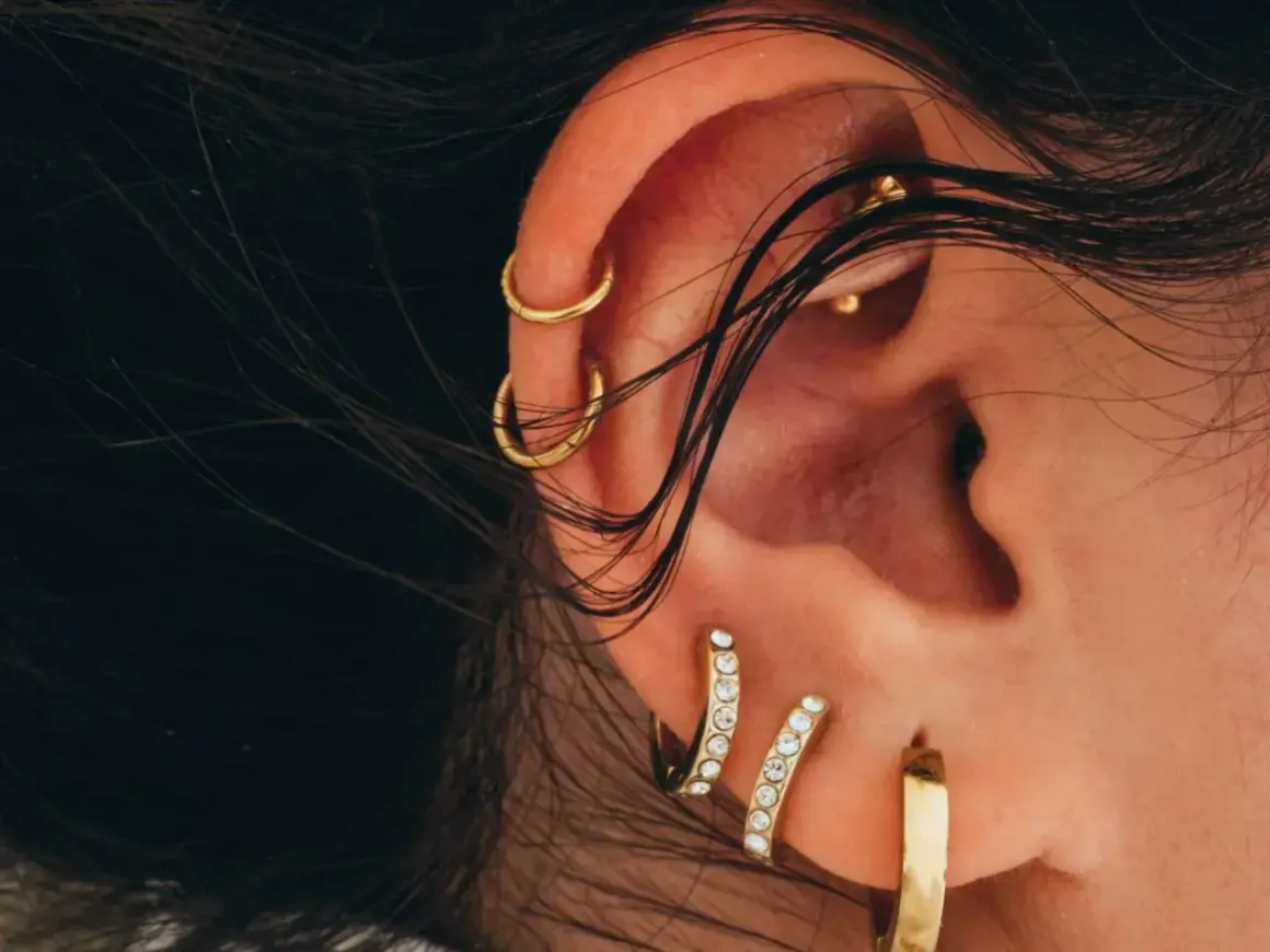 Złote kolczyki zdobią ucho, w tym modne conch przekłucie. Włosy delikatnie opadają na skórę.