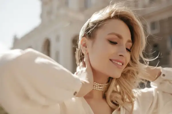 Uśmiechnięta blondynka z perłową opaską i naszyjnikiem. Różne rozmiary łańcuszków podkreślają jej szyję.