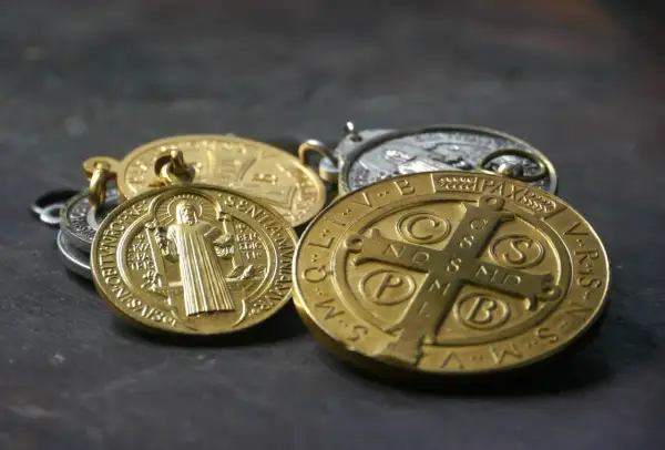 Kilka złotych i srebrnych medalików św. Benedykta leży na ciemnym tle.